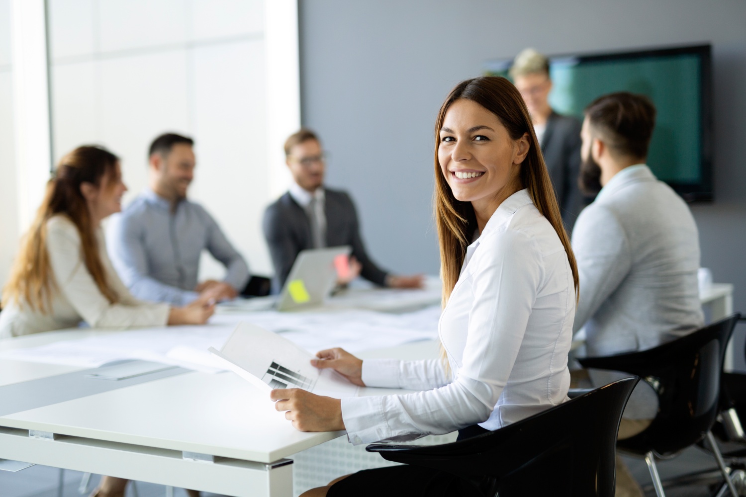 In einem hellen und modernen Büro sitzt eine lächelnde Frau mit Dokumenten in der Hand an einem Tisch, an dem weitere Personen an einer Besprechung teilnehmen.  Das Bild zeigt ein Besprechungszimmer mit einer Gruppe von etwa sieben Personen. Im Vordergrund sitzt eine Frau mit langen, braunen Haaren, die in einer weißen Bluse und einem schwarzen Rock gekleidet ist. Sie hält ein Dokument mit einem Diagramm in den Händen und lächelt in die Kamera. Hinter ihr sitzen weitere Personen an einem langen, weißen Tisch. Einige der Personen sind Männer in Anzügen oder Hemden, andere sind Frauen. Auf dem Tisch liegen Papiere und ein Laptop. Im Hintergrund ist eine große, graue Wand und ein dunkler Bildschirm zu sehen. Die Atmosphäre wirkt professionell und positiv. Das Bild vermittelt eine Stimmung von Zusammenarbeit und Erfolg.