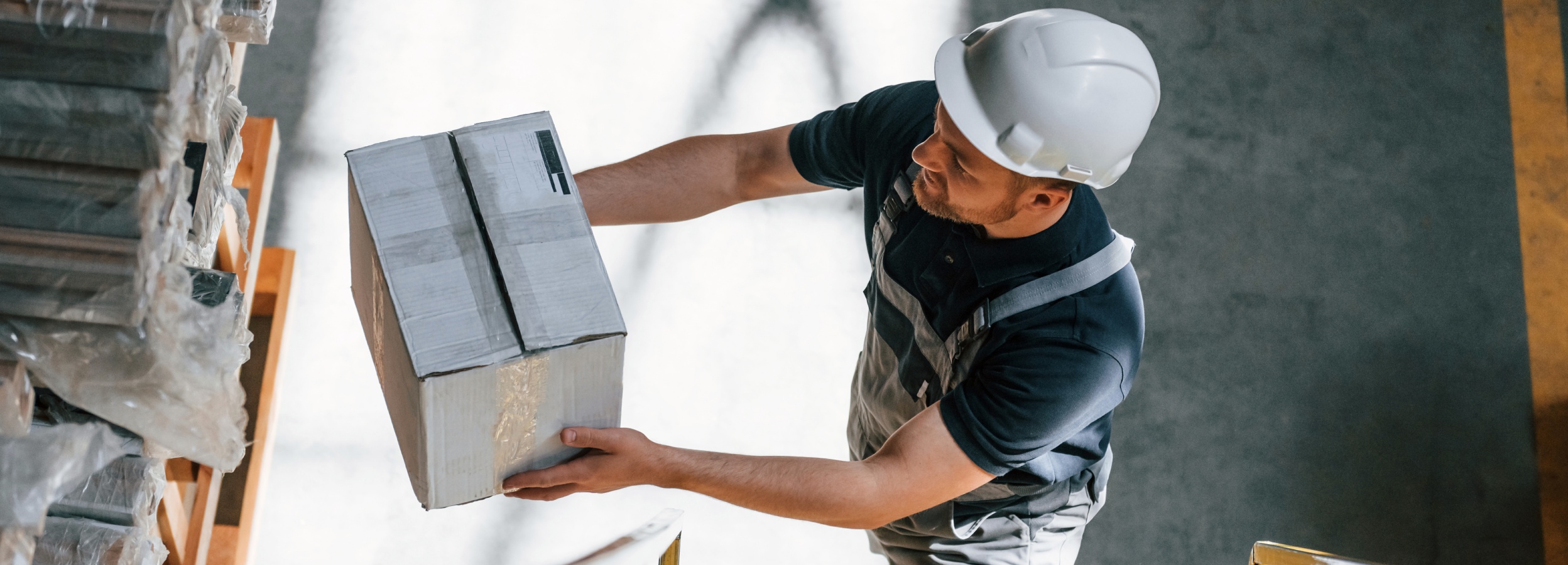 Blick von oben auf einen Lagerarbeiter, der ein Paket in ein Hochregal einräumt und einen Helm trägt.