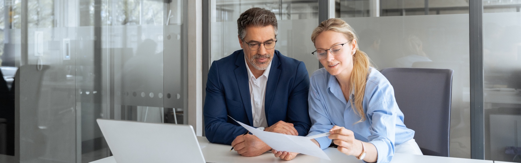Das Bild zeigt zwei Berufstätige, die in einem modernen Büro an einem Tisch sitzen und Dokumente besprechen.  Im Detail zeigt das Bild einen Mann und eine Frau, beide mit Brille, die an einem weißen Tisch sitzen. Der Mann trägt einen marineblauen Anzug über einem weißen Hemd und blickt auf die Dokumente, die die Frau in der Hand hält. Die Frau trägt ein hellblaues Hemd und weiße Hosen. Vor ihnen auf dem Tisch steht ein Laptop. Im Hintergrund ist ein Büroraum mit Glaswänden zu erkennen, der eine professionelle und moderne Atmosphäre vermittelt. Die Stimmung wirkt konzentriert und kooperativ.
